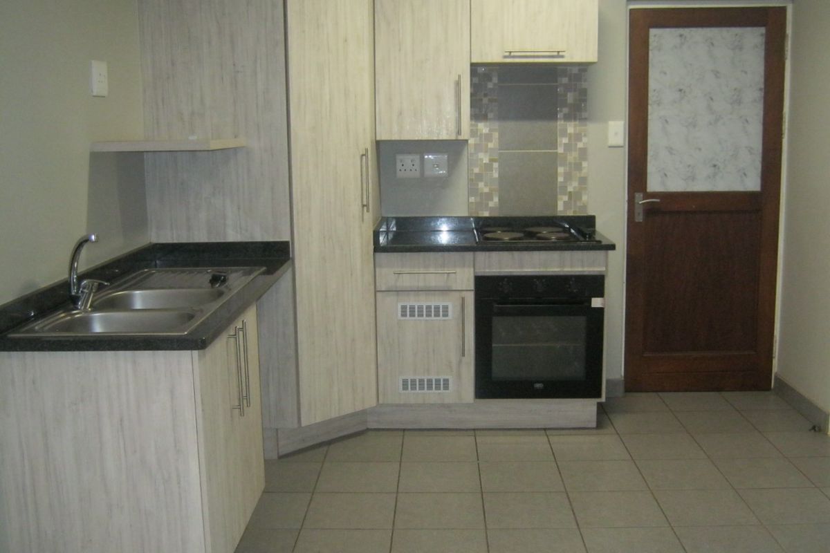 A compact, functional kitchen featuring light wood-grain cabinetry, dark laminate countertops, built-in oven and hob, and neutral tiled flooring.