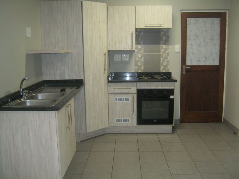A compact, functional kitchen featuring light wood-grain cabinetry, dark laminate countertops, built-in oven and hob, and neutral tiled flooring.
