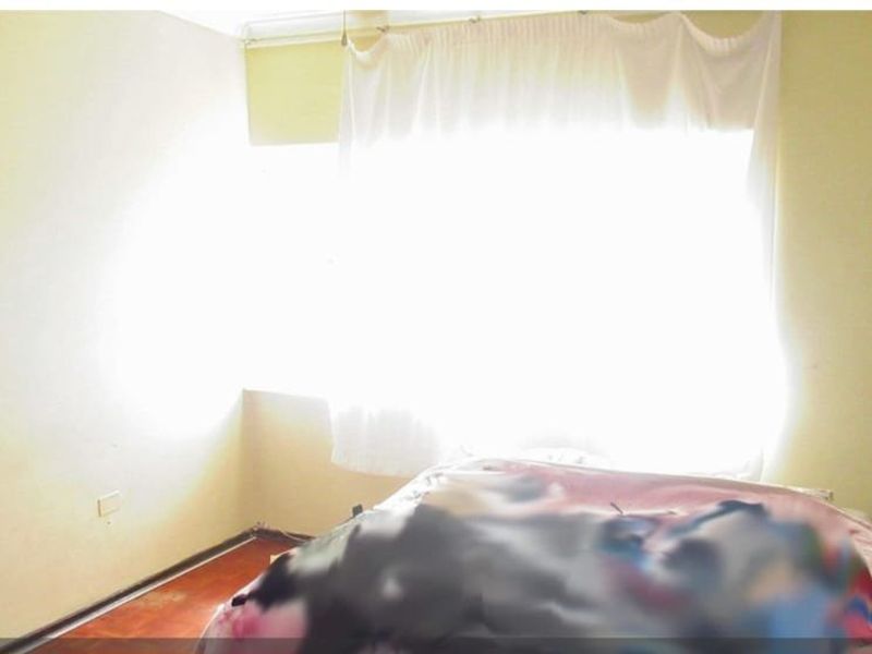 A bedroom featuring a large window with curtains and wooden flooring, currently showing signs of wear and tear.