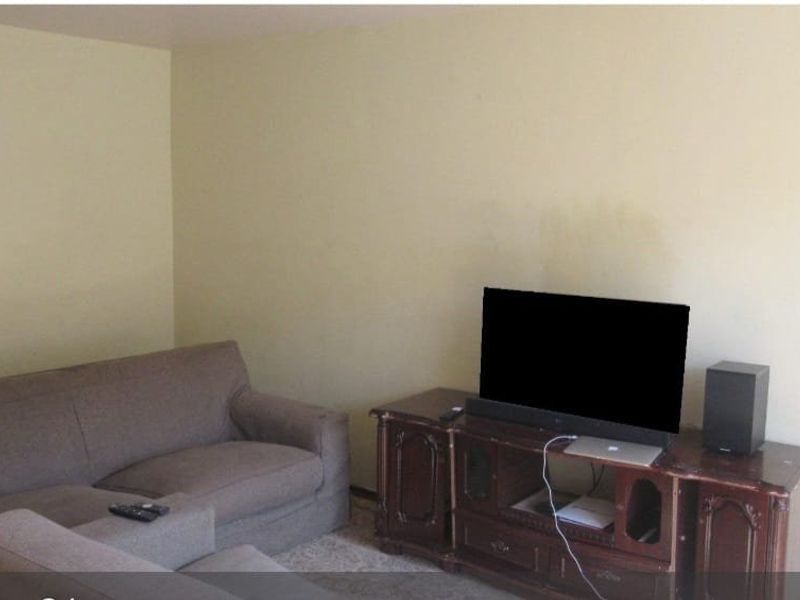 A living room area featuring plain walls and neutral carpeting, currently furnished with a sofa and media console.