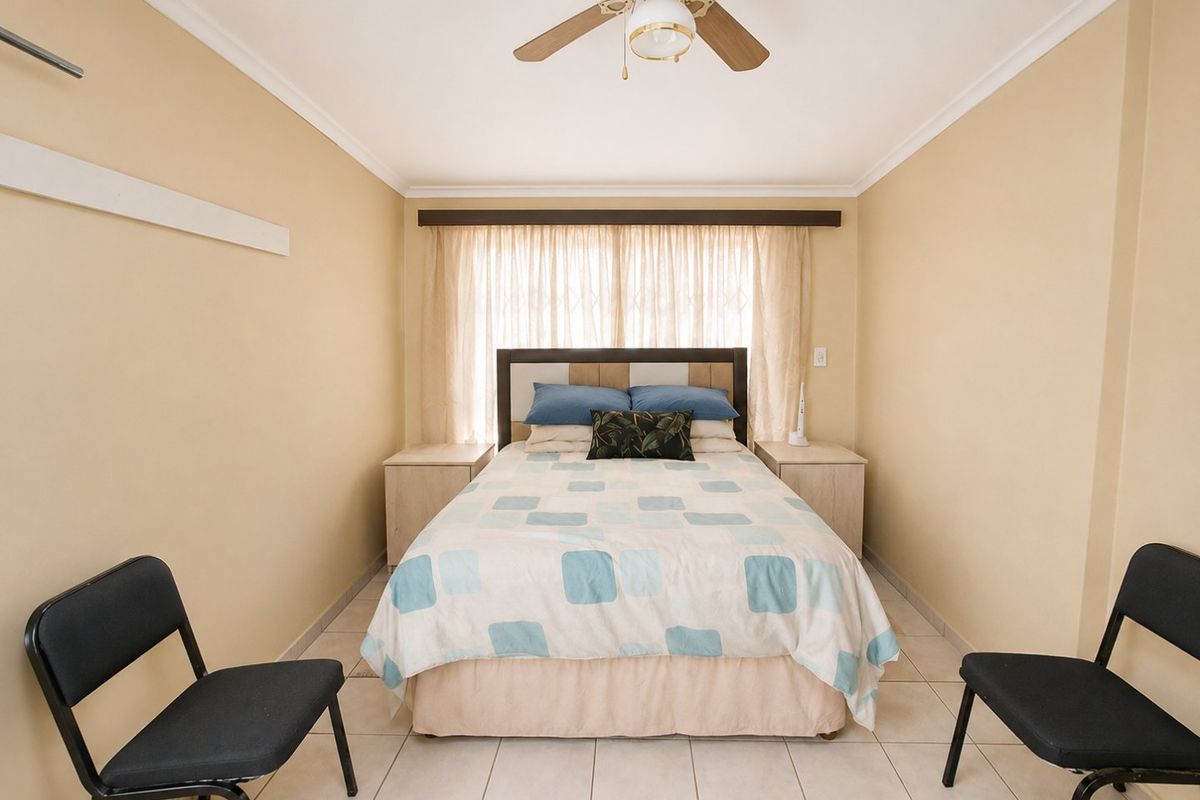 A functional bedroom featuring tiled flooring, a ceiling fan, and a window with curtain rails and security gate.