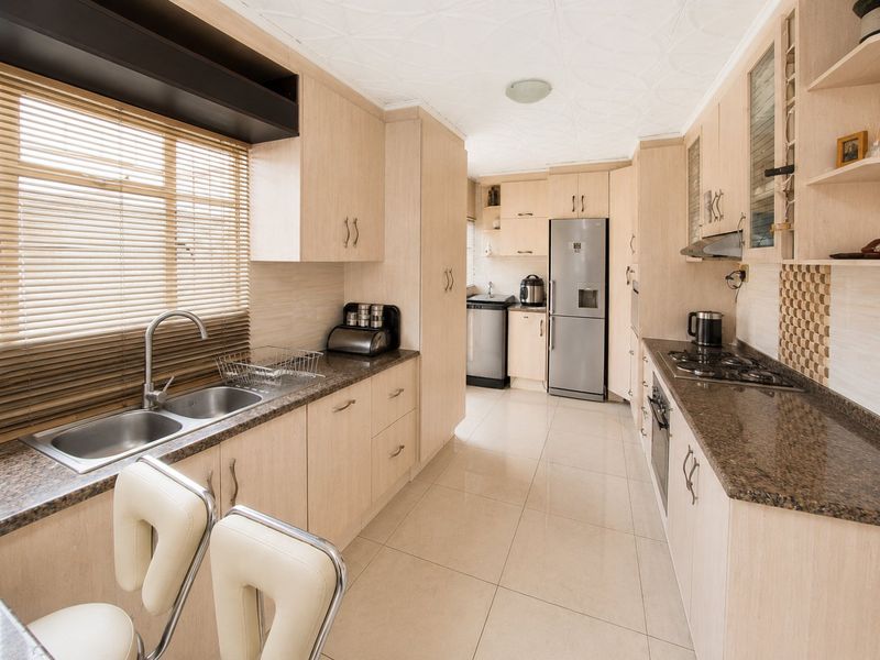 A functional galley-style kitchen featuring light wood cabinetry, granite countertops, a double basin sink, and integrated appliances with neutral tiled flooring.
