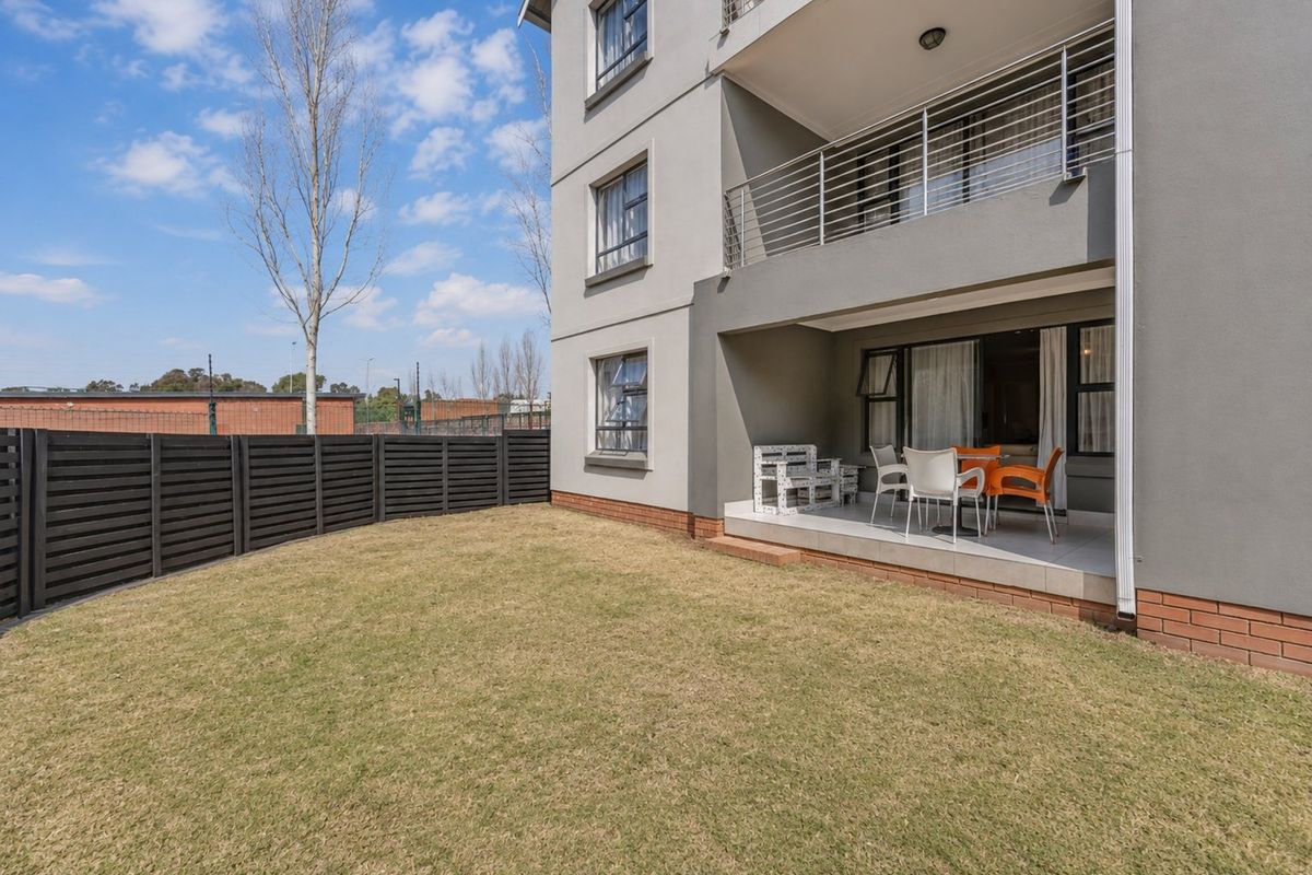 Rear view of the apartment building featuring a private ground-floor tiled patio, a small lawn area, and a secure perimeter fence.