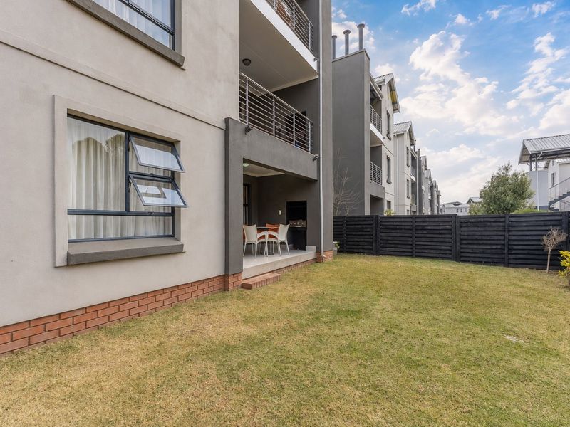 Rear view of the apartment building featuring a covered patio with a built-in braai, metal balcony railings, and a private garden area.