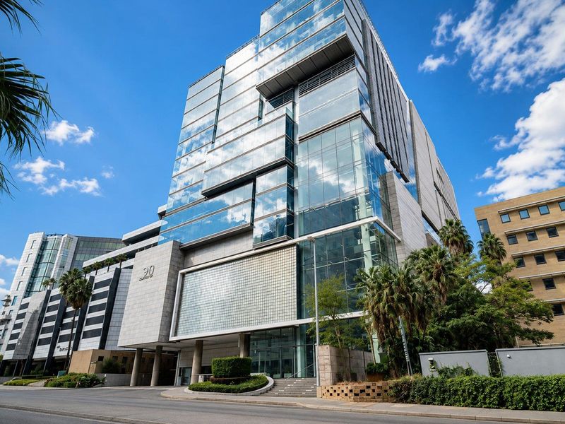 Modern commercial office building featuring a prominent glass curtain wall facade, stone cladding, and professional landscaping at the street-level entrance.