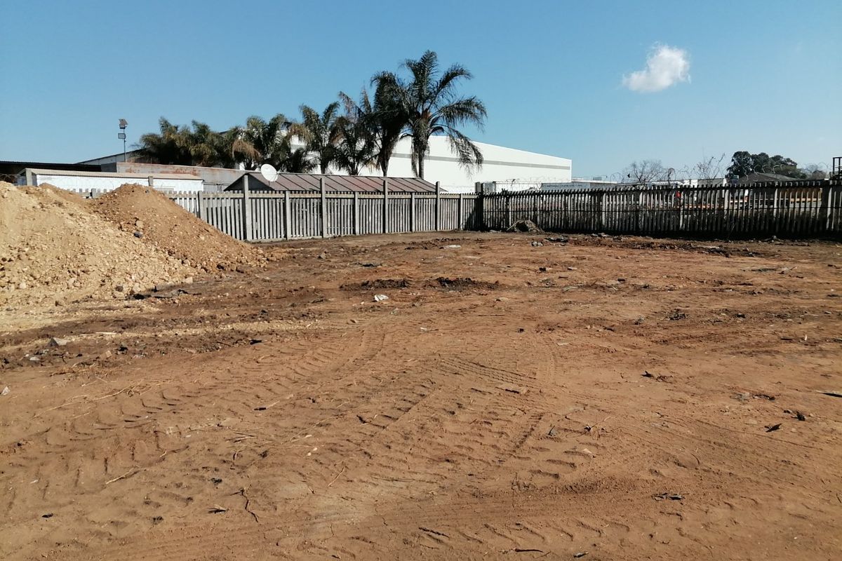 A vacant residential plot featuring cleared earth, perimeter fencing, and surrounding industrial-style structures.
