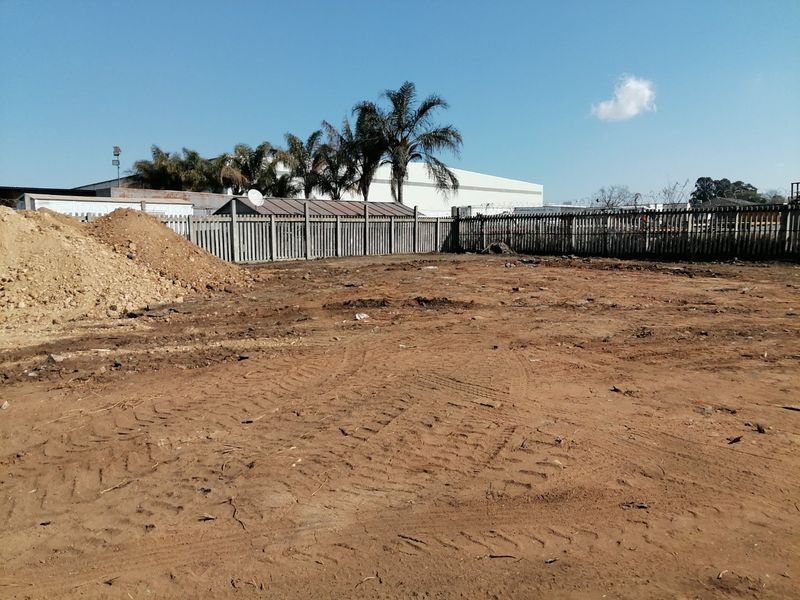 A vacant residential plot featuring cleared earth, perimeter fencing, and surrounding industrial-style structures.