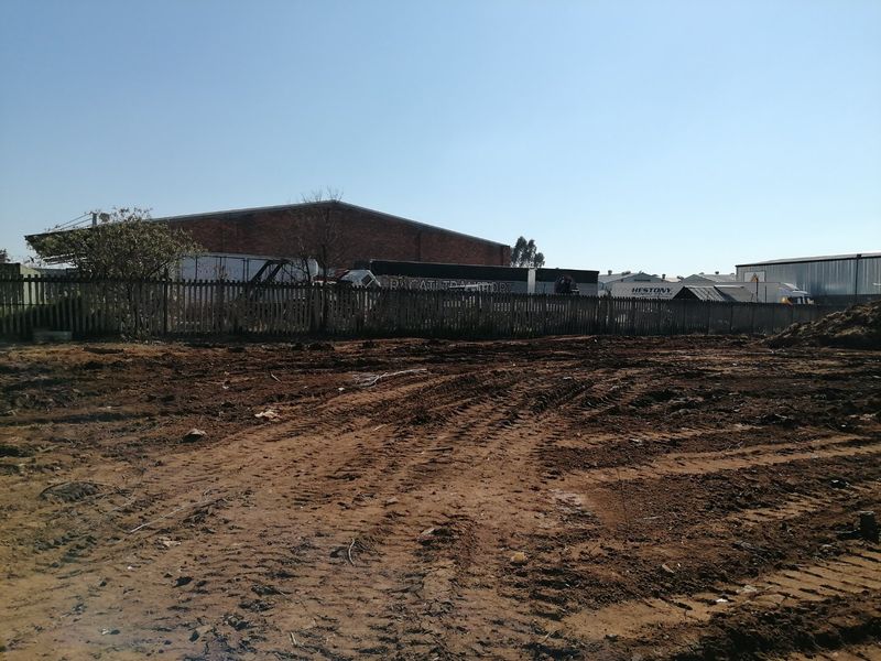 A view of a vacant residential plot featuring cleared earth and a wooden boundary fence, with industrial buildings visible in the background.