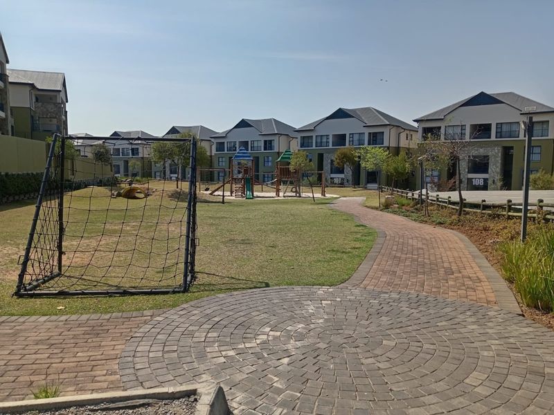 A well-maintained communal garden area featuring a paved walkway, landscaped lawn, and playground equipment within a residential apartment complex.