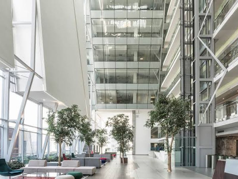 Spacious modern commercial atrium featuring a high glass curtain wall, steel structural elements, and a central reception area with integrated elevator shafts.