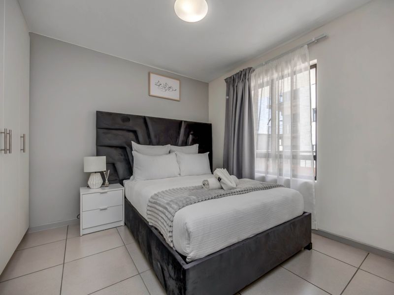 A well-maintained bedroom featuring neutral tiled flooring, a built-in wardrobe, and natural light through a window with curtain tracks.