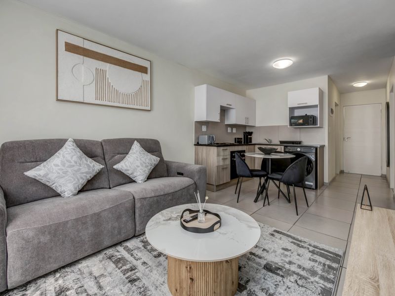 A modern open-plan living area featuring a compact kitchen with built-in cabinetry, integrated appliances, and neutral tiled flooring throughout.