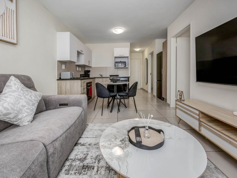 Modern open-plan living area featuring a compact kitchen with built-in cabinetry, integrated appliances, and neutral tiled flooring throughout.