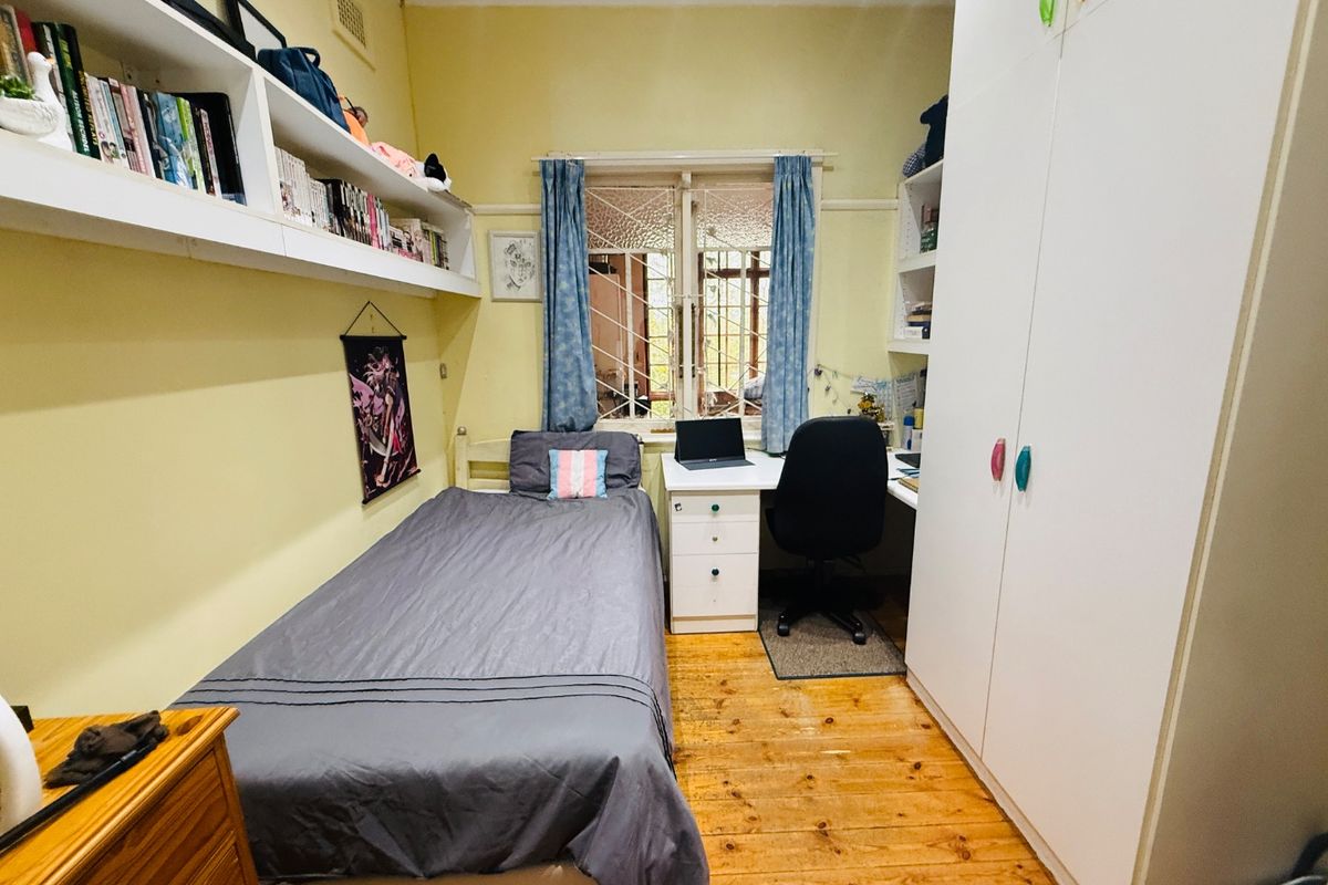 A functional bedroom featuring timber flooring, built-in wardrobe storage, floating shelving, and a window with security bars.