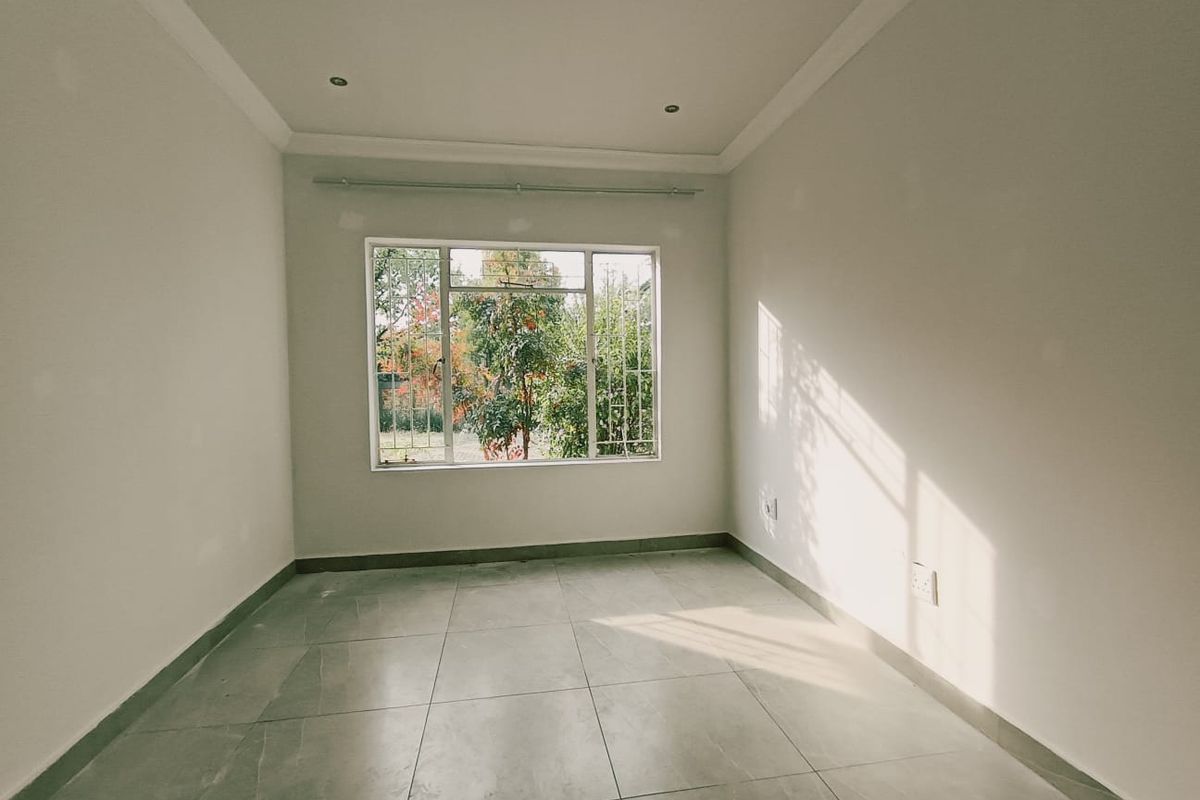 A bright, empty bedroom featuring neutral tiled flooring, recessed downlighting, and a large window with security bars overlooking the garden.