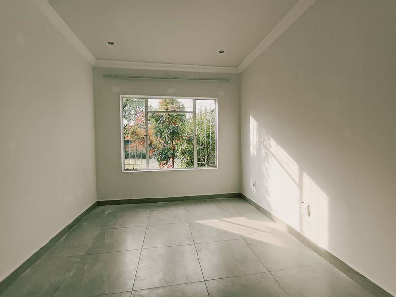 A bright, empty bedroom featuring neutral tiled flooring, recessed downlighting, and a large window with security bars overlooking the garden.