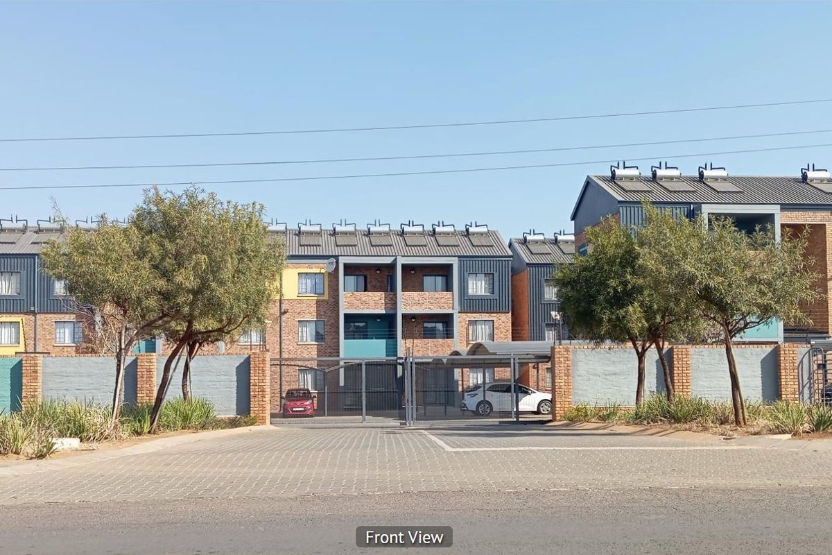 Front view of a multi-story apartment complex featuring brick facade, metal roofing with solar water heaters, and a secure gated entrance.