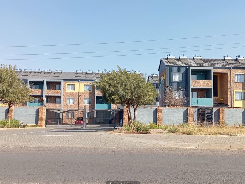 Front view of a multi-story apartment complex featuring brick facades, balconies, and solar water heating systems, secured by a perimeter wall and gate.