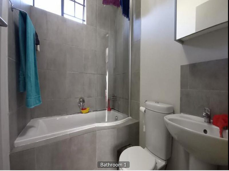 A functional bathroom featuring a bathtub with a glass shower screen, a pedestal basin, and a toilet with modern grey wall tiling.