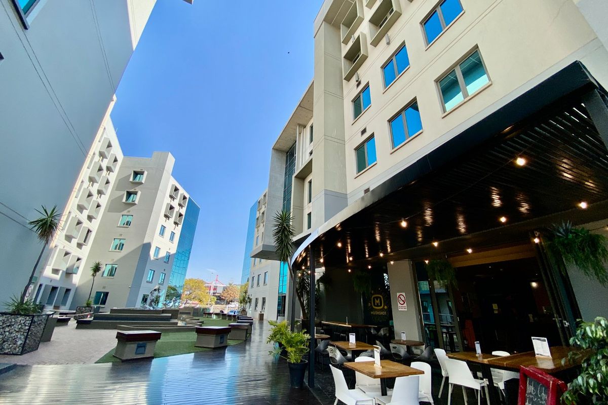 A modern commercial outdoor patio area featuring timber decking, overhead lighting, and access to a landscaped courtyard between multi-story office or hotel buildings.