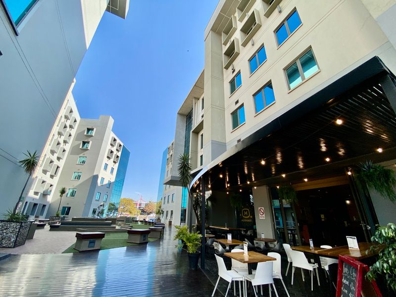 A modern commercial outdoor patio area featuring timber decking, overhead lighting, and access to a landscaped courtyard between multi-story office or hotel buildings.