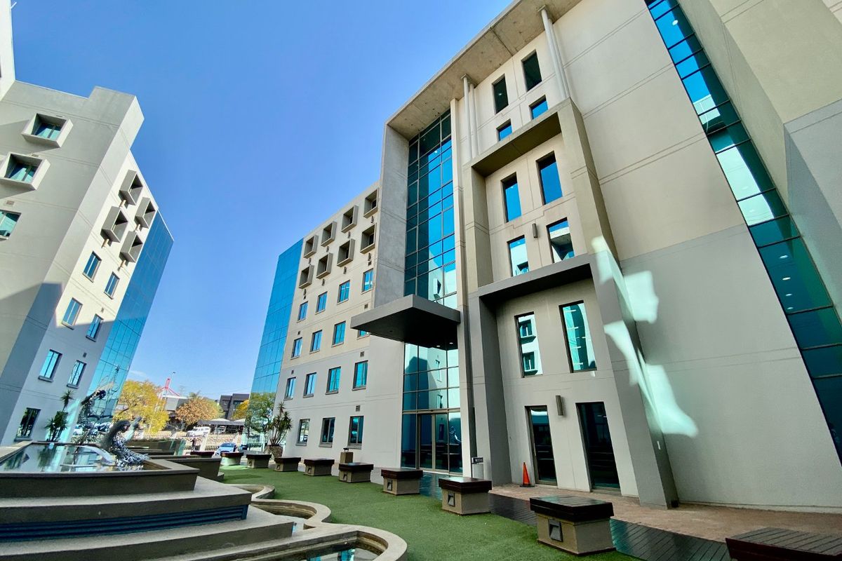 Modern commercial building exterior featuring a glass facade, concrete structure, landscaped courtyard with a water feature, and integrated outdoor seating areas.