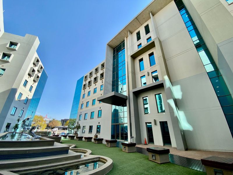 Modern commercial building exterior featuring a glass facade, concrete structure, landscaped courtyard with a water feature, and integrated outdoor seating areas.
