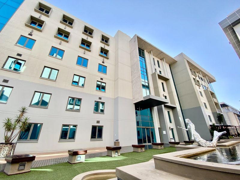 Modern multi-story commercial building facade featuring a glass entrance, landscaped courtyard with a water feature, and integrated outdoor seating areas.