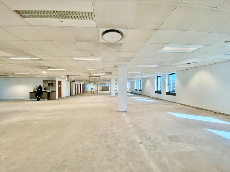 Expansive open-plan commercial office space featuring a suspended ceiling, recessed lighting, structural columns, and a small built-in kitchenette area.