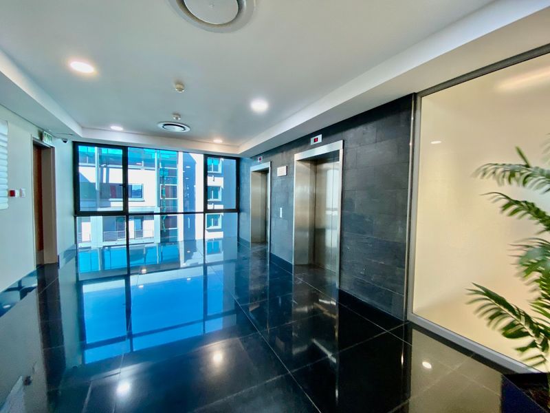 Modern commercial building lobby featuring polished dark tile flooring, stainless steel elevator banks, and a stone feature wall with large windows.