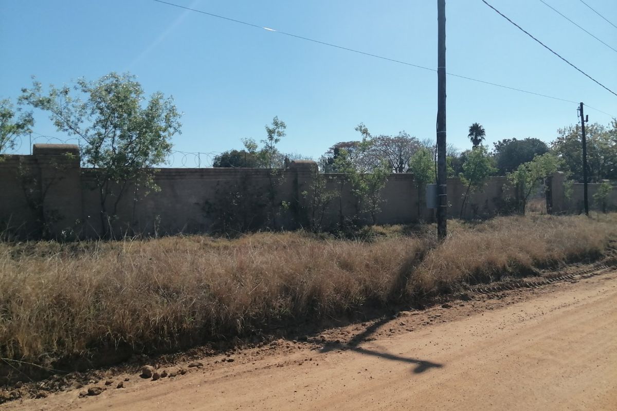 Street view of a vacant residential plot featuring a perimeter brick wall with razor wire and adjacent utility infrastructure along an unpaved road.