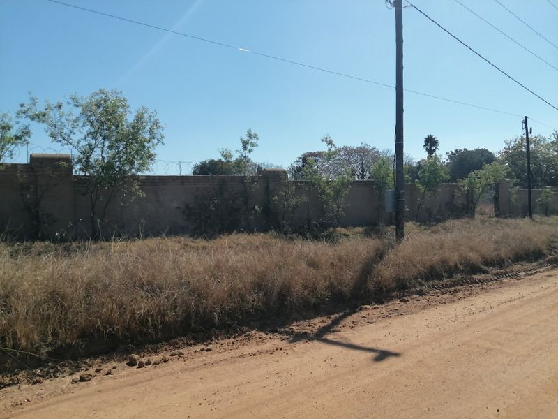 Street view of a vacant residential plot featuring a perimeter brick wall with razor wire and adjacent utility infrastructure along an unpaved road.