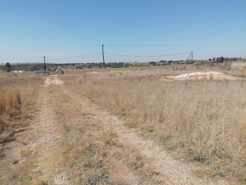 A vacant residential plot featuring a dirt driveway, perimeter walling, and overhead power lines, set within an open, undeveloped landscape.