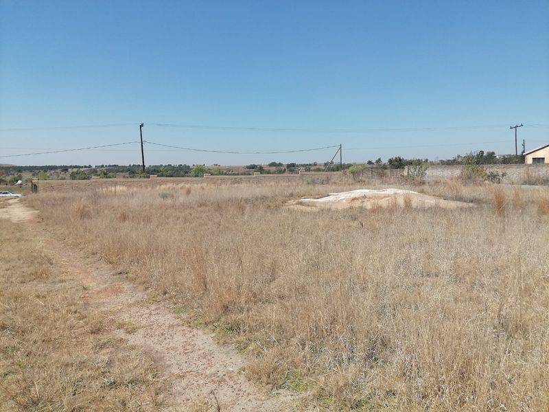 A vacant residential plot featuring dry grass, perimeter walling, and overhead power lines, offering a clear space for future development.