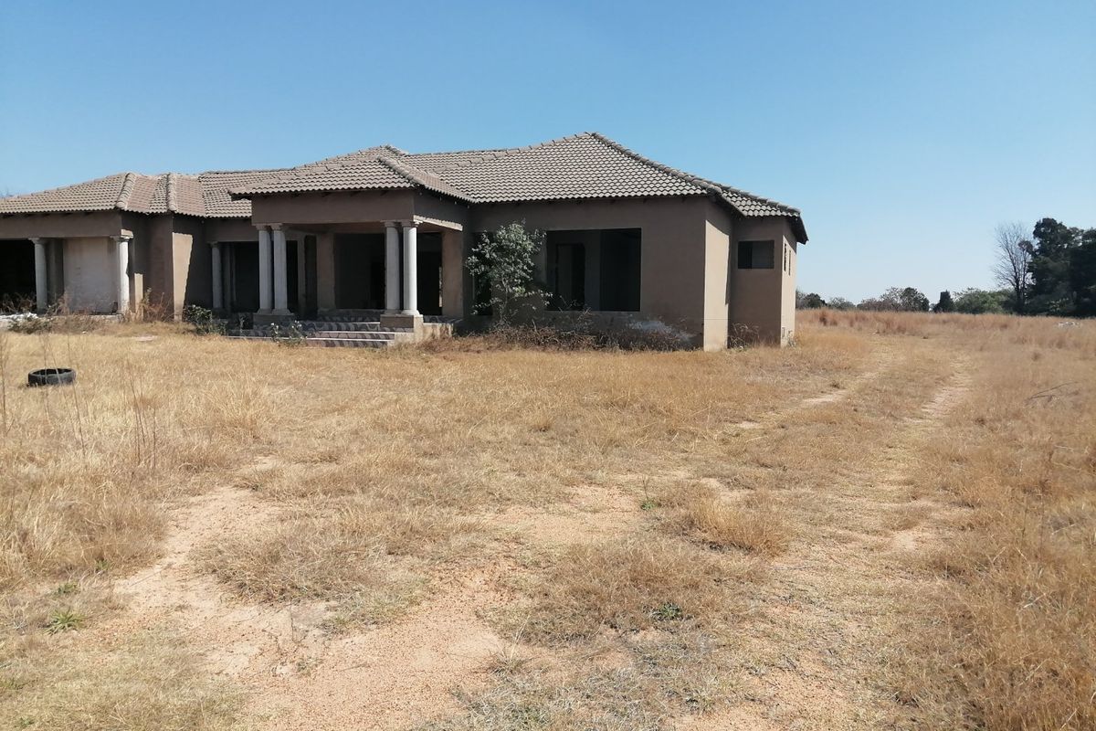 An unfinished single-story residential property featuring a tiled roof and concrete entrance columns, set on a large, undeveloped plot of land.