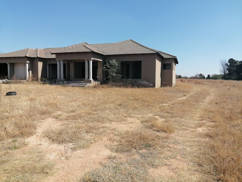 An unfinished single-story residential property featuring a tiled roof and concrete entrance columns, set on a large, undeveloped plot of land.