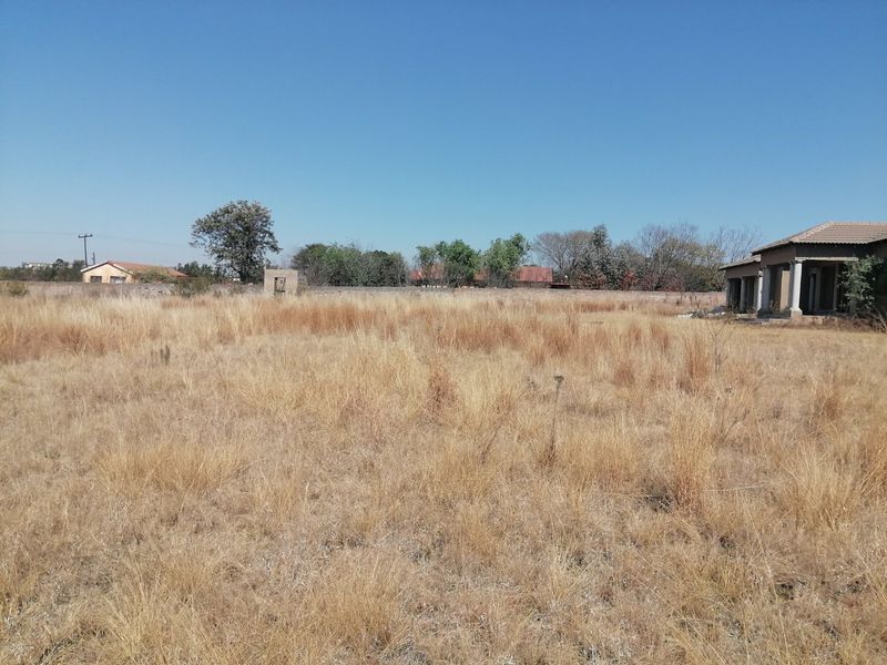 A wide view of a large, undeveloped plot of land featuring dry grass, a perimeter wall, and a partial view of a residential structure.