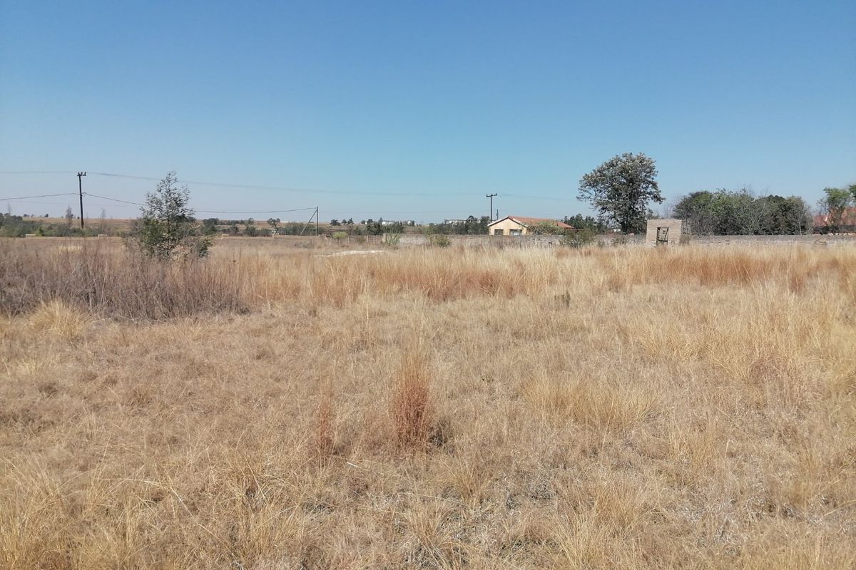 A large, undeveloped plot of land featuring dry grass, a boundary wall, and a small outbuilding structure in the distance.