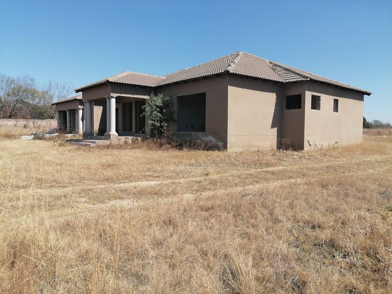 An unfinished residential property structure featuring a tiled roof and concrete columns, set on a large, undeveloped plot of land.