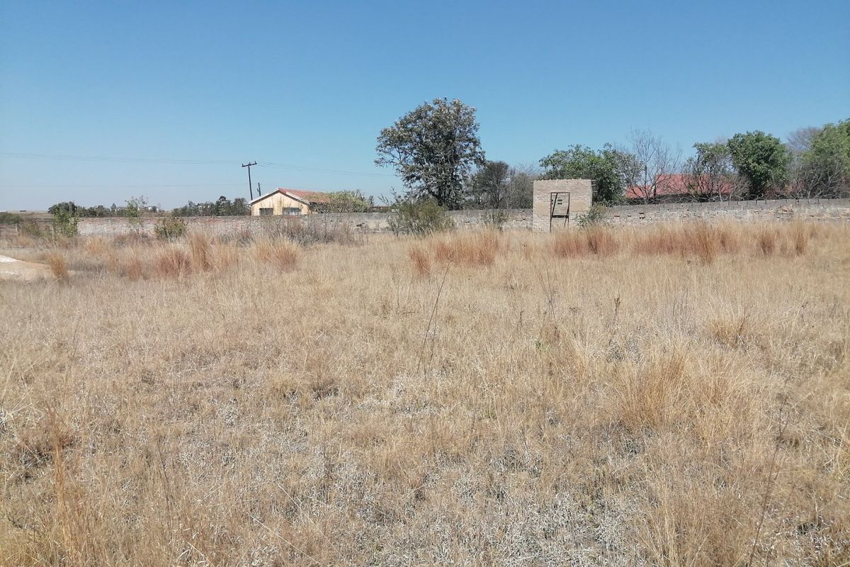 A view of a vacant residential plot featuring dry grass, a perimeter wall, and a small brick structure in the background.