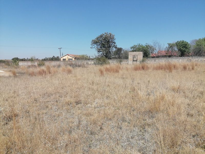 A view of a vacant residential plot featuring dry grass, a perimeter wall, and a small brick structure in the background.