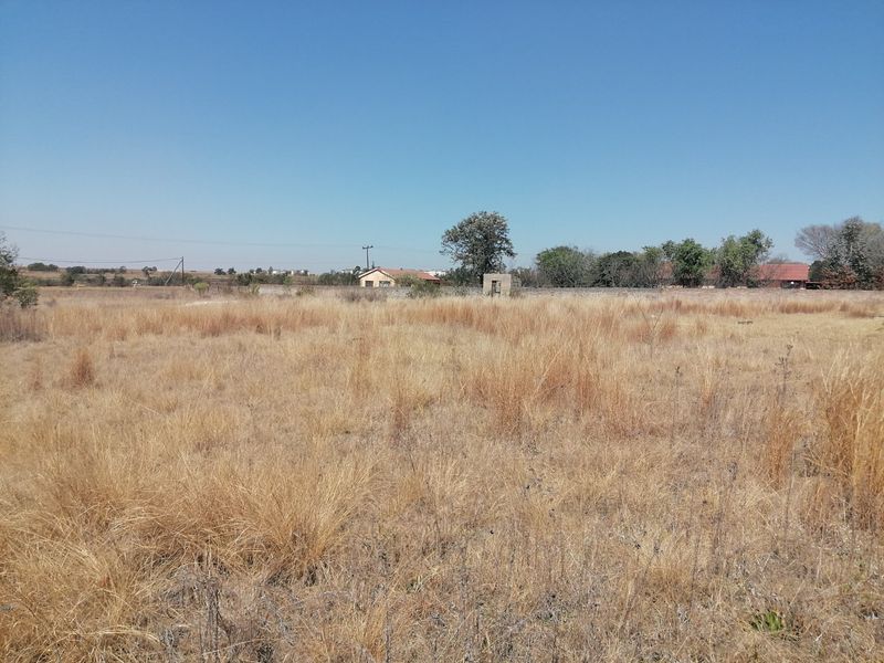 A wide-angle view of a vacant residential plot featuring dry grass and a distant boundary wall under a clear blue sky.