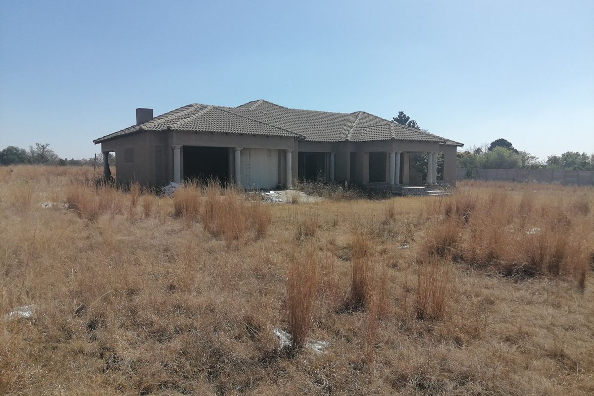An unfinished residential property structure situated on a large, overgrown vacant plot of land.