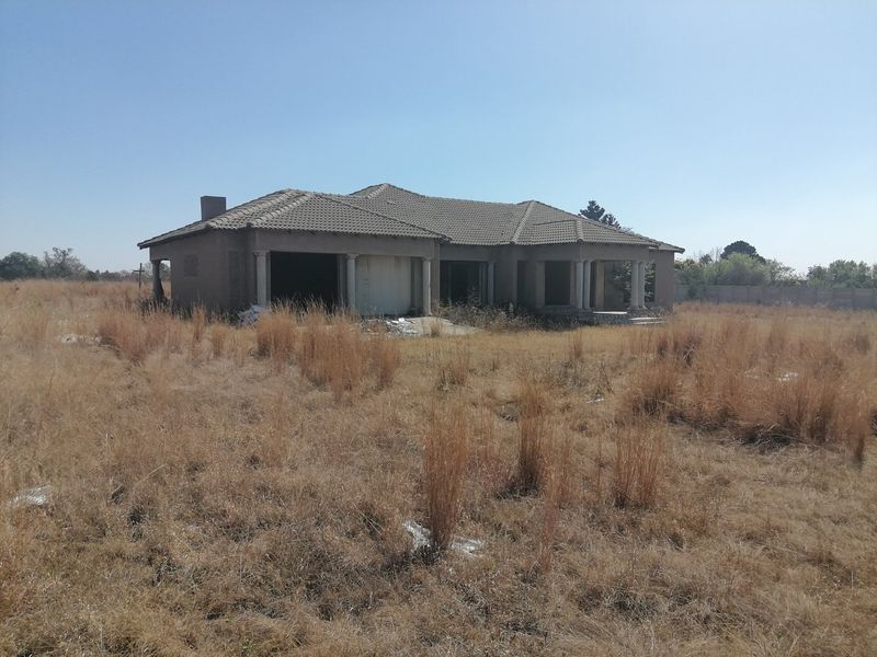 An unfinished residential property structure situated on a large, overgrown vacant plot of land.