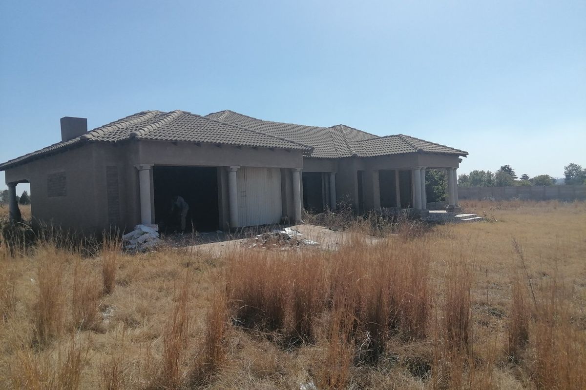 An unfinished residential property featuring a tiled roof, concrete columns, and a garage, situated on a large plot of vacant land.