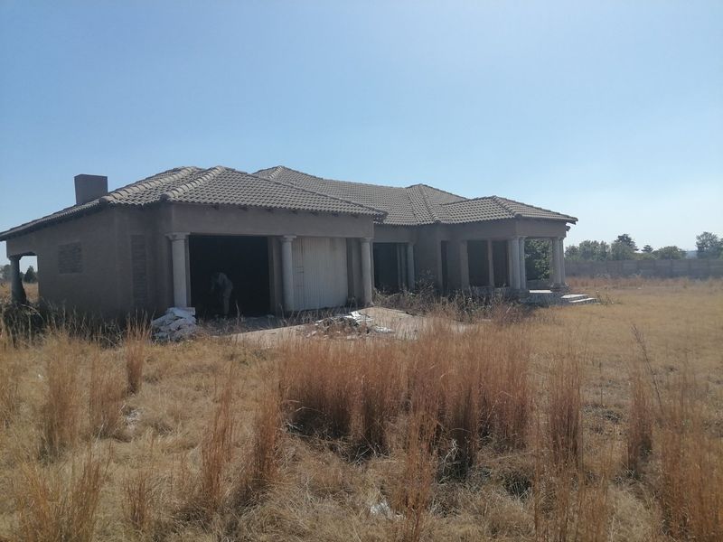 An unfinished residential property featuring a tiled roof, concrete columns, and a garage, situated on a large plot of vacant land.