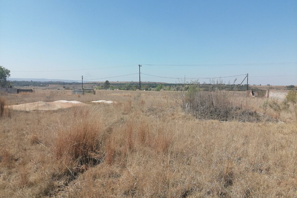 A view of a vacant residential plot featuring dry grass, perimeter walling, and overhead utility lines under a clear sky.