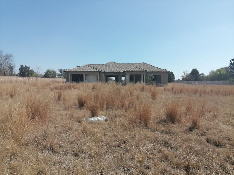 A view of an incomplete residential structure situated on a large, undeveloped plot of land with dry, overgrown grass.
