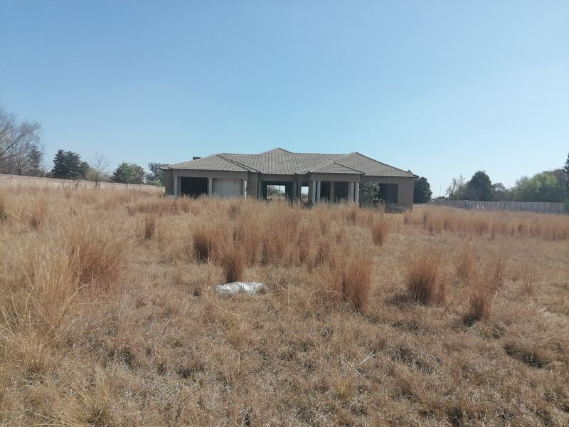A view of an incomplete residential structure situated on a large, undeveloped plot of land with dry grass and perimeter fencing.