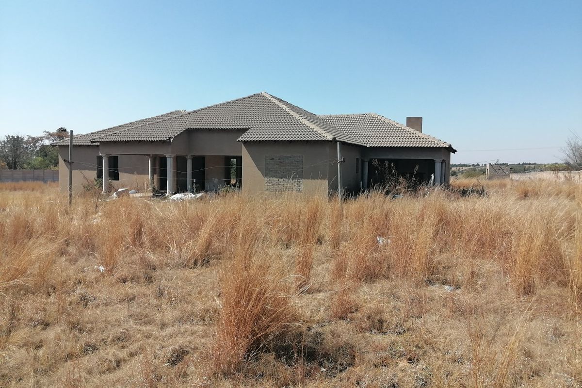 An unfinished residential property structure set on a large plot of land with overgrown grass and clear signs of incomplete construction.